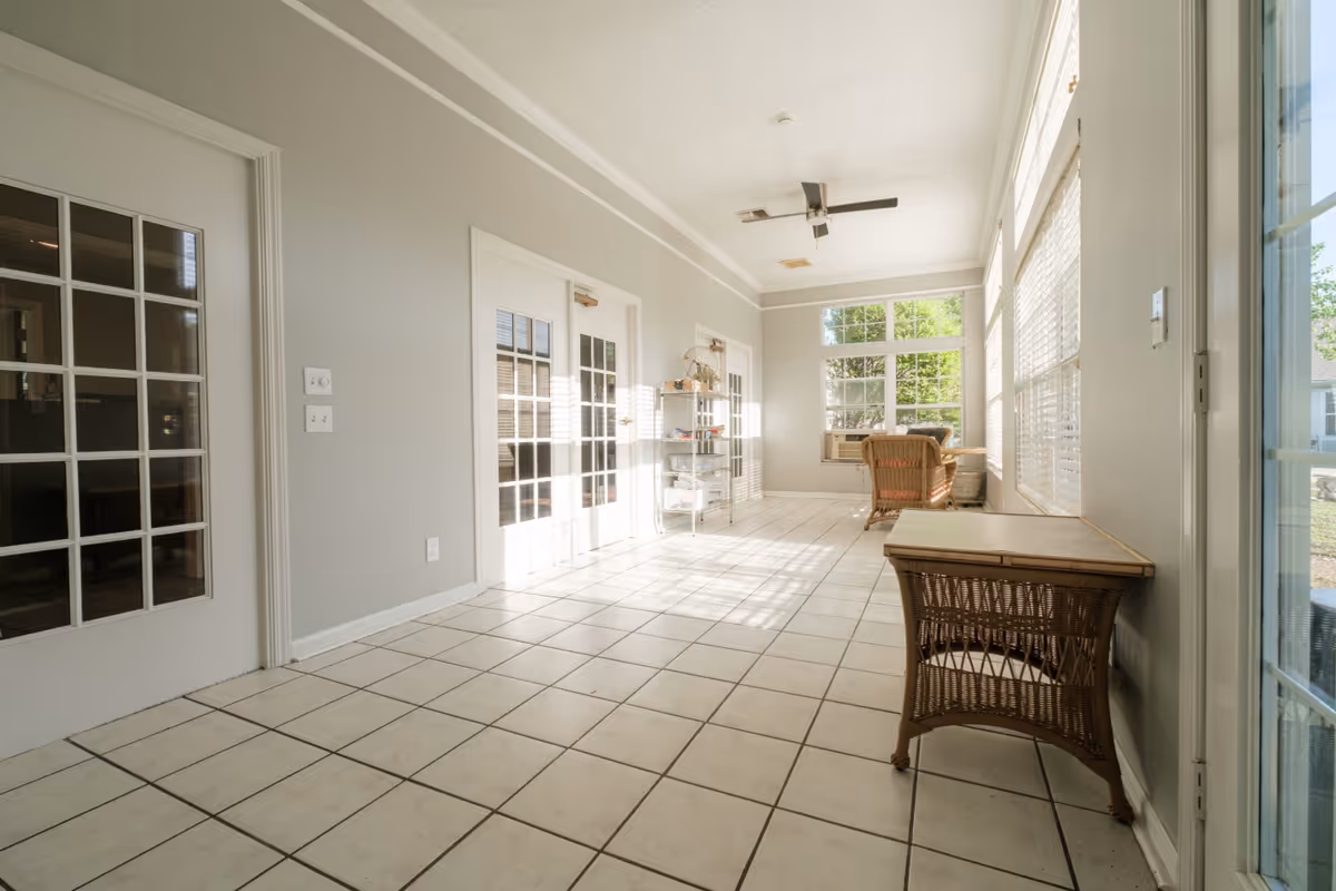A bright sunroom with large windows on two sides allowing natural light to fill the space. The room has white tiled flooring, light gray walls, and white trim. There are two wicker chairs and a wicker table, a metal shelving unit with some items on it, and a ceiling fan with light fixtures. French doors with glass panes lead to another room.