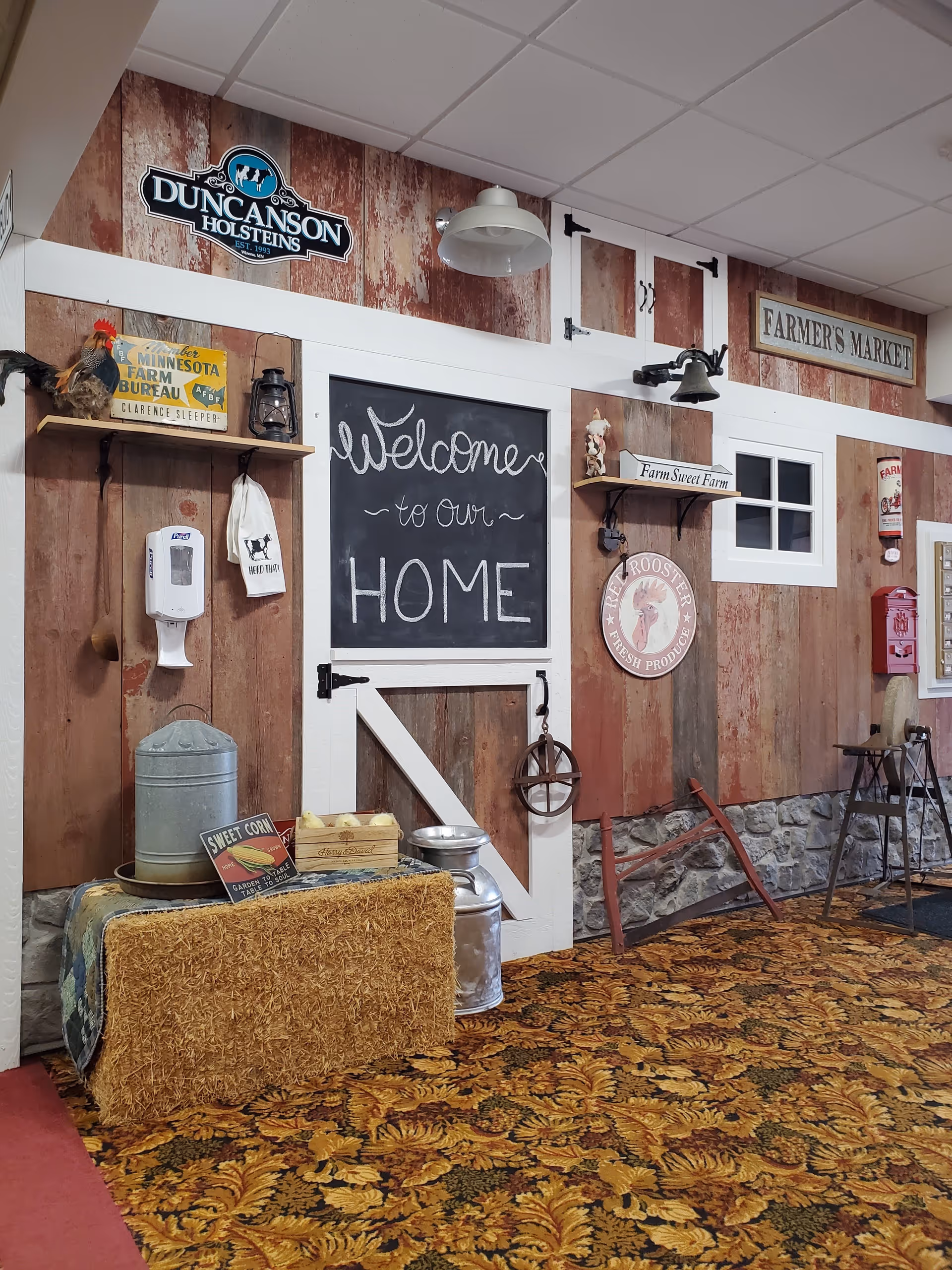 Interior wall decorated with rustic farm-themed items including a chalkboard sign that says 'Welcome to our HOME', a metal milk can, a bale of hay, a wooden crate with yellow chicks, vintage signs, a rooster figurine, and a patterned carpet with autumn leaves design.