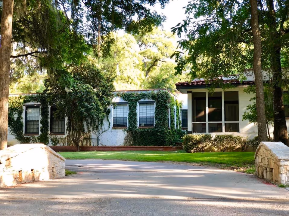 Exterior view of a white building partially covered with green ivy and surrounded by trees and bushes. There is a paved driveway leading up to the building with a small stone bridge on each side. The building has multiple windows and a covered porch area.