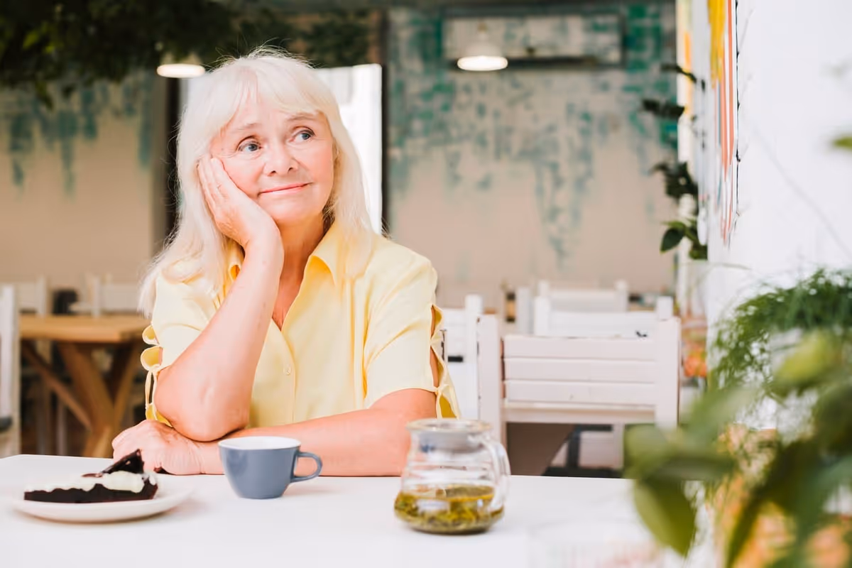 An elderly woman with white hair wearing a yellow shirt sits at a table in a bright dining area. She rests her head on her hand and looks thoughtfully to the side. On the table in front of her are a cup, a glass teapot with tea, and a plate with a slice of chocolate cake. The background shows white chairs and tables with some greenery on the walls.