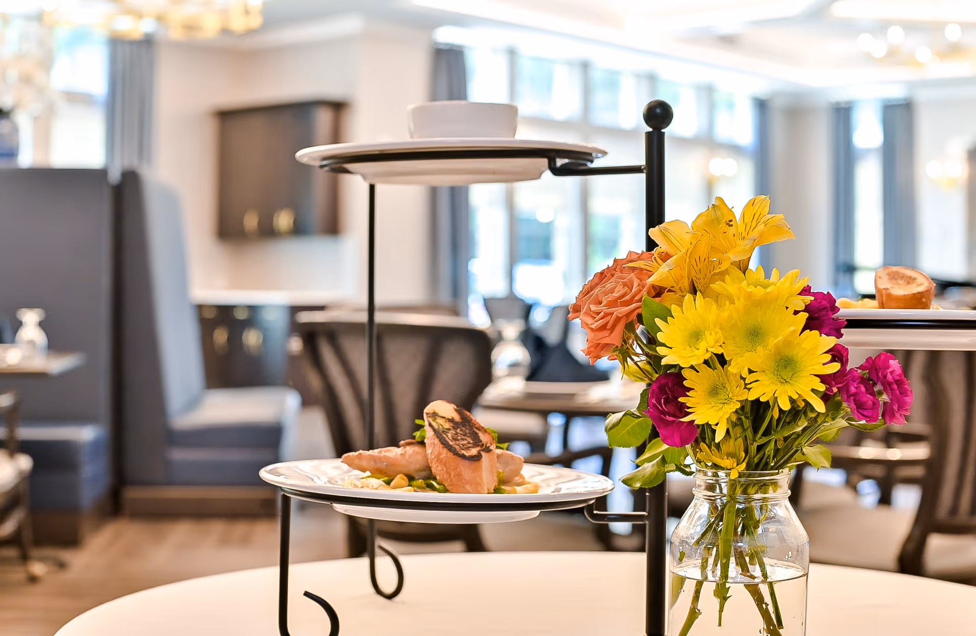 A close-up view of a dining table with a two-tiered serving stand holding plates of food and a cup, next to a glass jar filled with a colorful bouquet of yellow, pink, and orange flowers. The background shows a softly focused dining area with chairs, tables, and large windows letting in natural light.