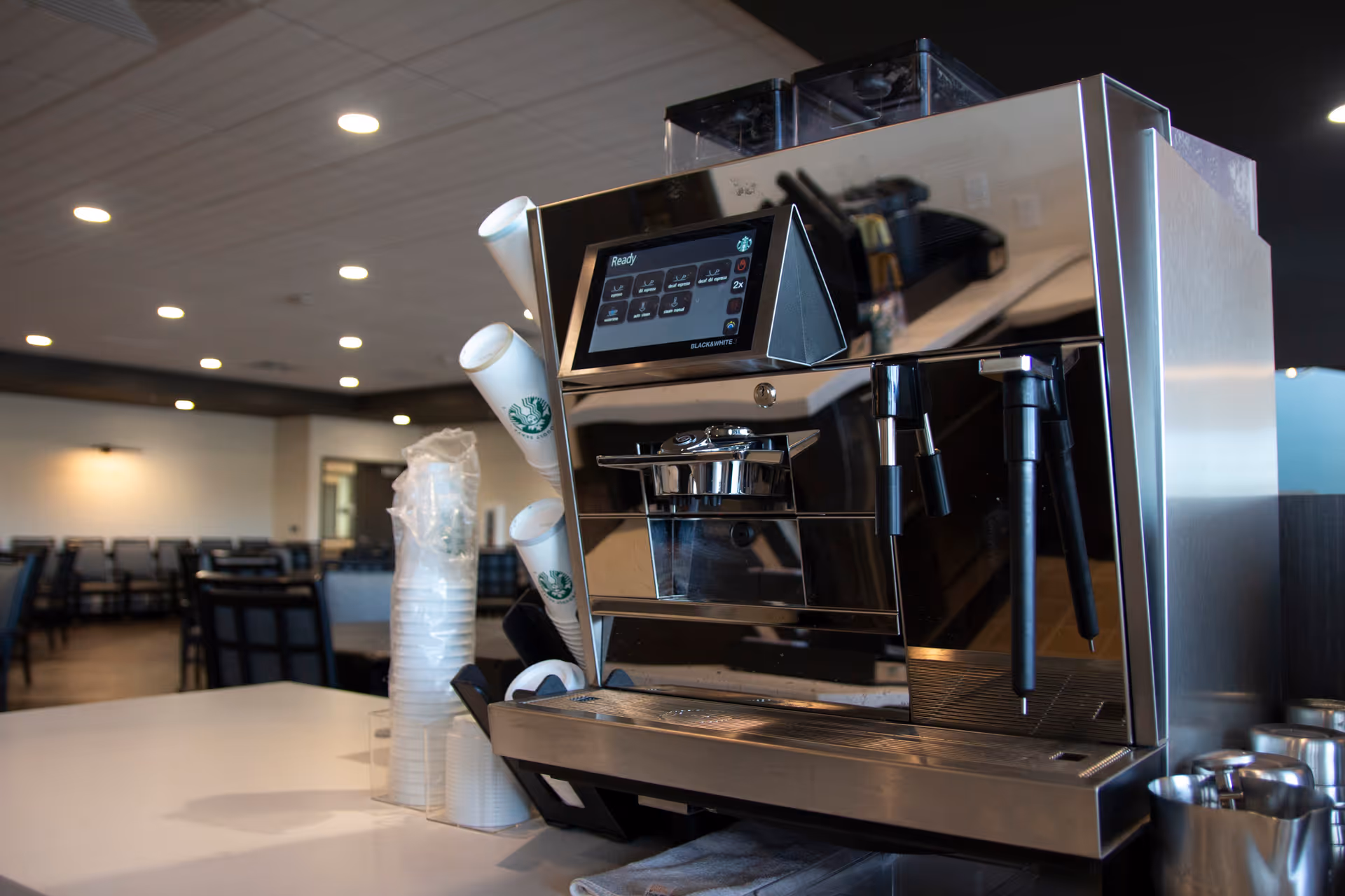 Commercial espresso machine and stacked disposable cups on a counter in a dining area with tables and chairs in the background.