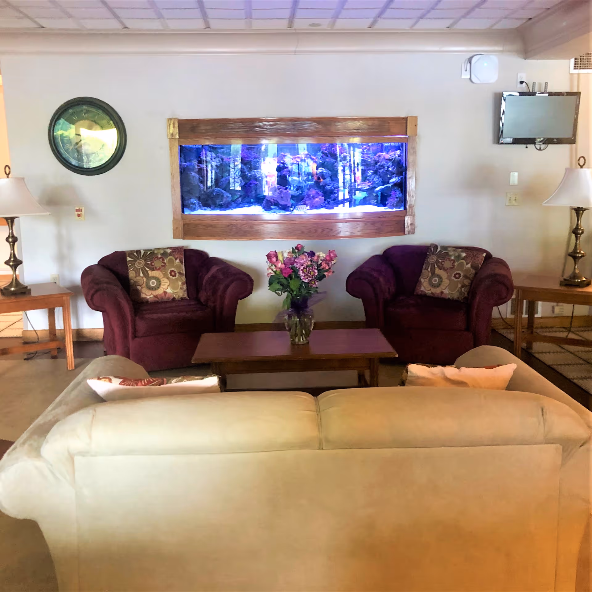 A cozy living room area with a beige sofa facing two burgundy armchairs with floral cushions. Between the armchairs is a wooden coffee table with a vase of flowers. On the wall behind the armchairs is a large built-in aquarium with colorful fish and rocks, flanked by two side tables with lamps. A round wall clock is on the left side of the wall, and a small flat-screen TV is mounted on the right side.