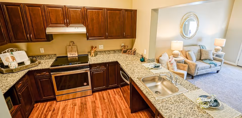 A modern kitchen with dark wooden cabinets, granite countertops, a stainless steel oven, and a sink. The kitchen opens up to a living area with a beige sofa, a chair, two lamps on side tables, and a round mirror on the wall.