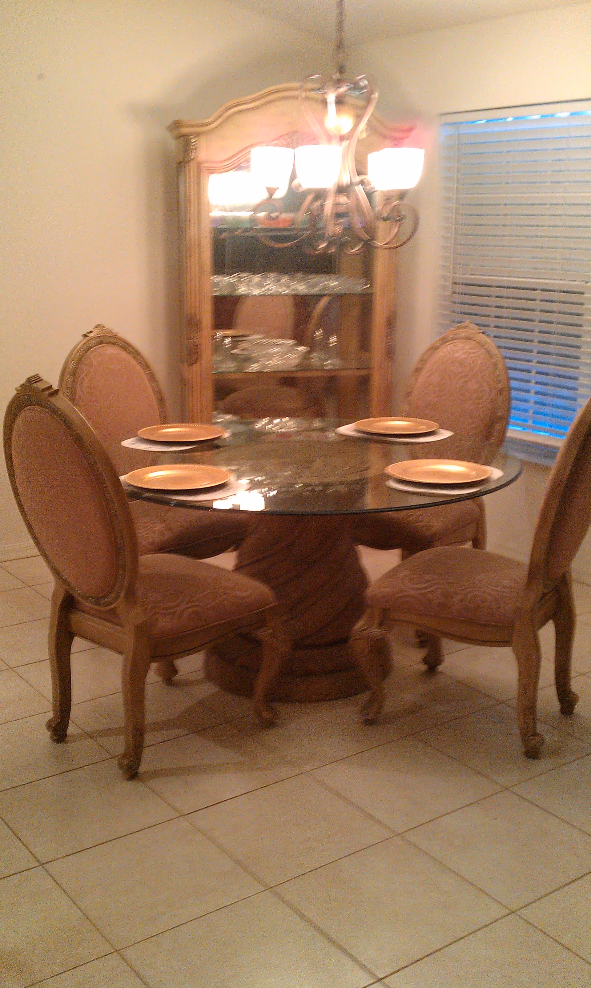 Round glass-top dining table with four upholstered chairs, place settings, a chandelier, and a china cabinet in a tiled room.