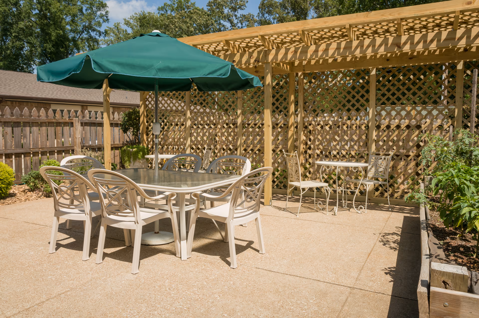 Outdoor patio with a glass-top dining table, several plastic chairs and a green umbrella under a wooden pergola and lattice fence.