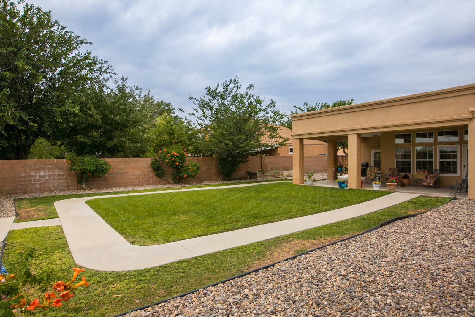 A well-maintained outdoor garden area with a green lawn bordered by a concrete walkway. There are trees and flowering plants along a brick wall fence. A covered patio with seating and potted plants is attached to a beige building under a cloudy sky.