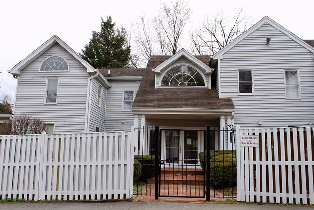 Exterior view of AlfredHouse Elder Care, Inc. facility with white siding, gate, and brick steps.