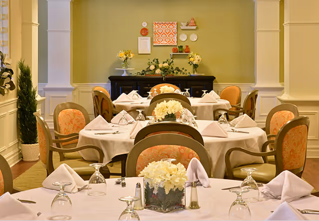 Dining room with round tables set with white tablecloths, folded napkins, glassware, floral centerpieces, and upholstered chairs against a pale green wall.