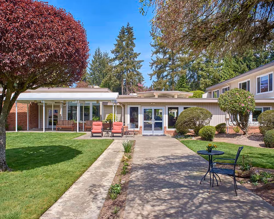 Outdoor view of Sagebrook Senior Living at Bellevue showing a walkway leading to the building entrance. The area is landscaped with green grass, trimmed bushes, a tree with red leaves, and outdoor seating including two red cushioned chairs and a small table with a chair. The sky is clear and blue.