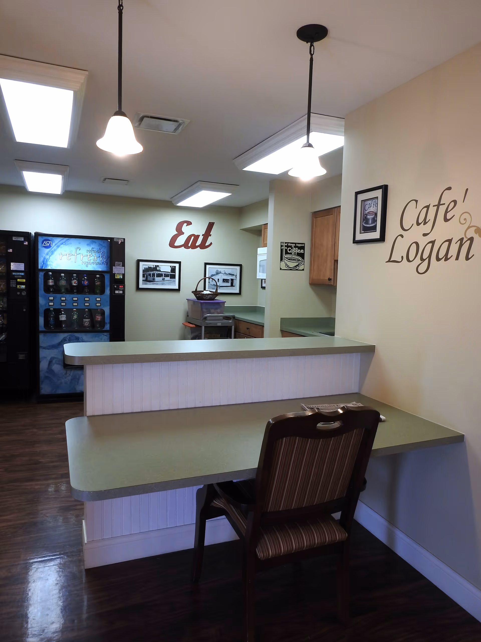 Interior view of a small cafe area named 'Cafe Logan' with a counter and a single chair. The walls have decorative text saying 'Eat' and 'Cafe Logan'. There are vending machines in the background and framed pictures on the wall. The lighting consists of ceiling lights and hanging pendant lamps.