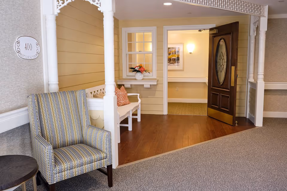 Interior entryway styled vignette with a patterned armchair, white bench with pillows, wood flooring and an open decorative wooden door.