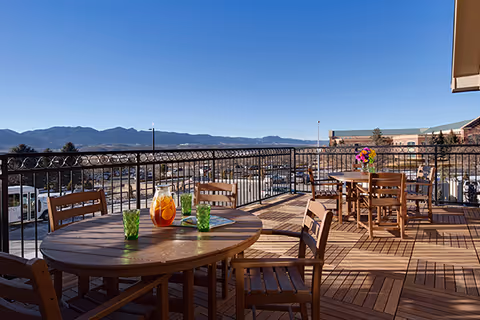 Sunny outdoor terrace with wooden dining tables and chairs, a pitcher and glasses on a table, and mountains in the background.