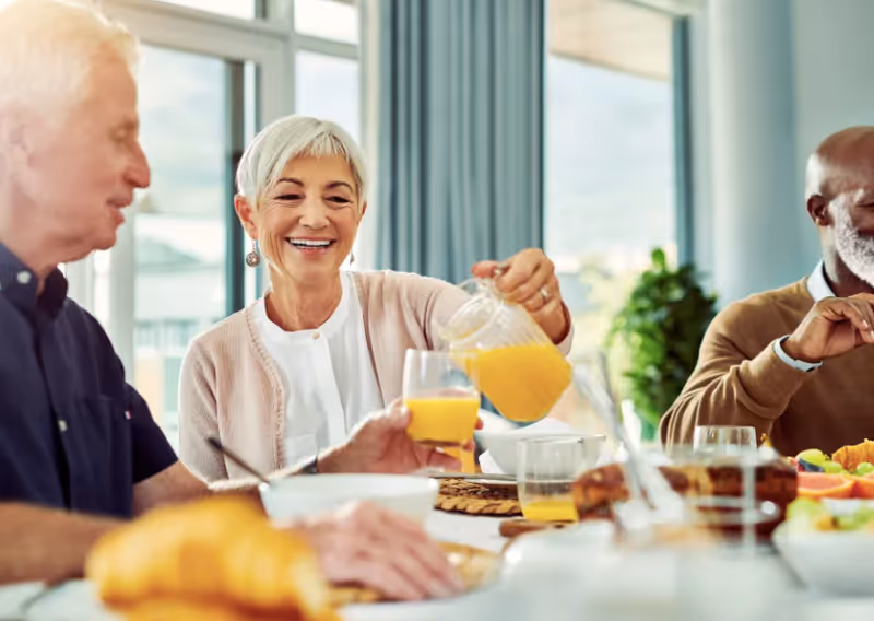 Three older adults seated at a table in a bright dining area, with a woman pouring orange juice into a glass.