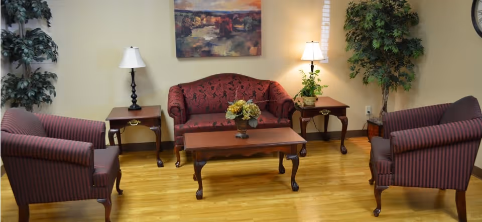 A cozy living room area with a red patterned loveseat, two matching armchairs with red and black stripes, a wooden coffee table with a flower arrangement, two wooden side tables each with a lamp, and two large potted plants. A colorful landscape painting hangs on the wall above the loveseat.