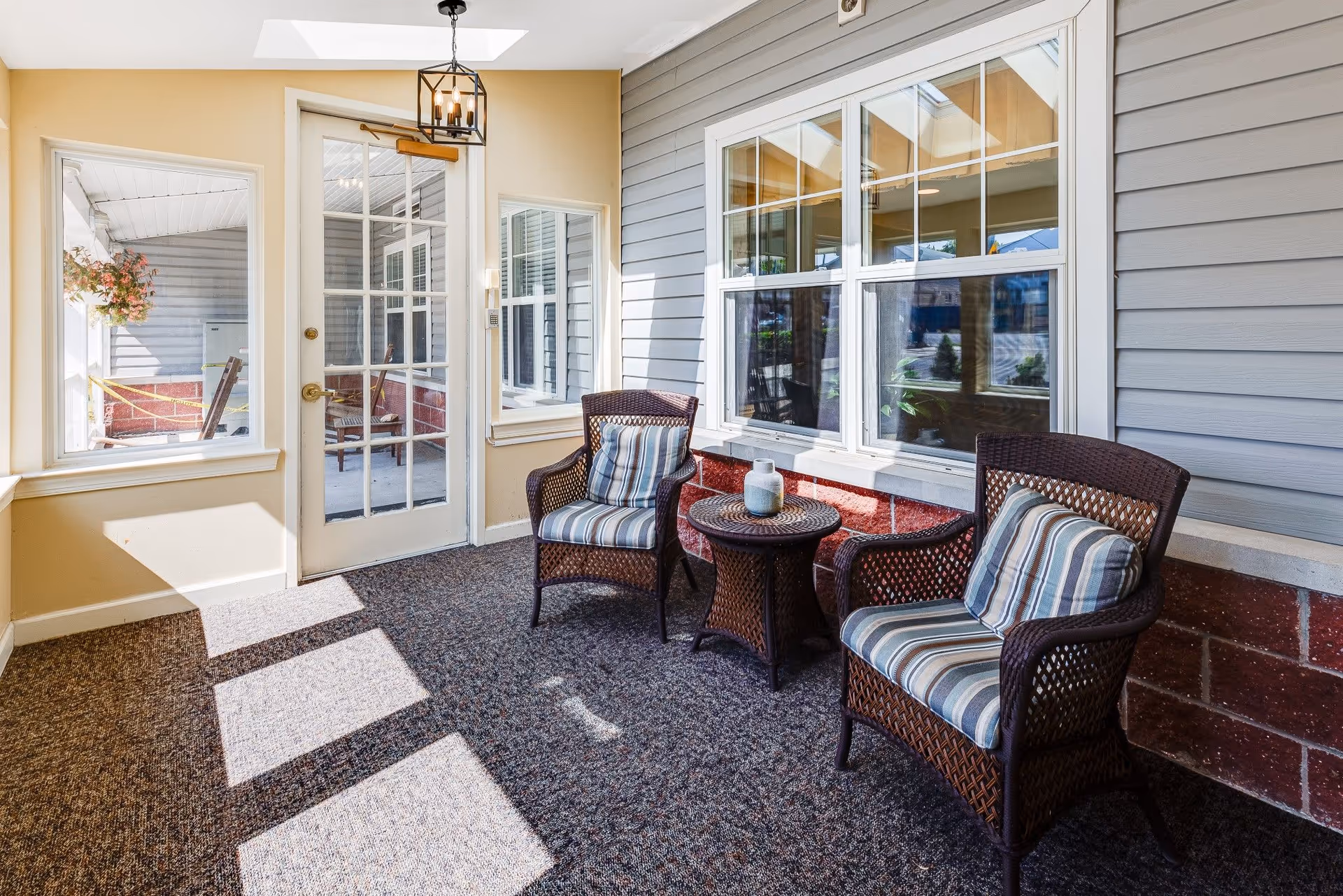 Sunlit enclosed porch area with two wicker chairs featuring striped cushions and a small round wicker table between them, a glass door with multiple panes, beige walls, and windows looking out to an outdoor space.