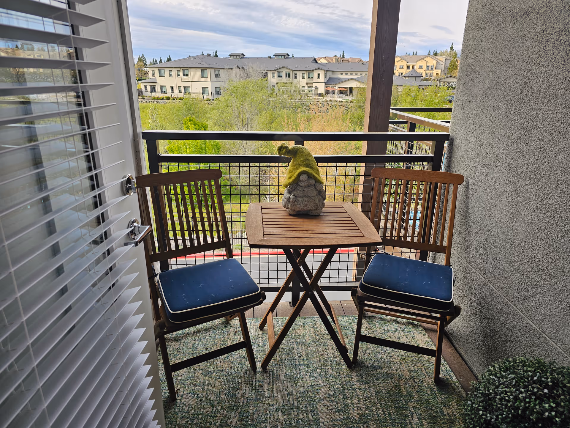 Small balcony with two wooden chairs and a folding table holding a gnome figurine, overlooking greenery and neighboring buildings.