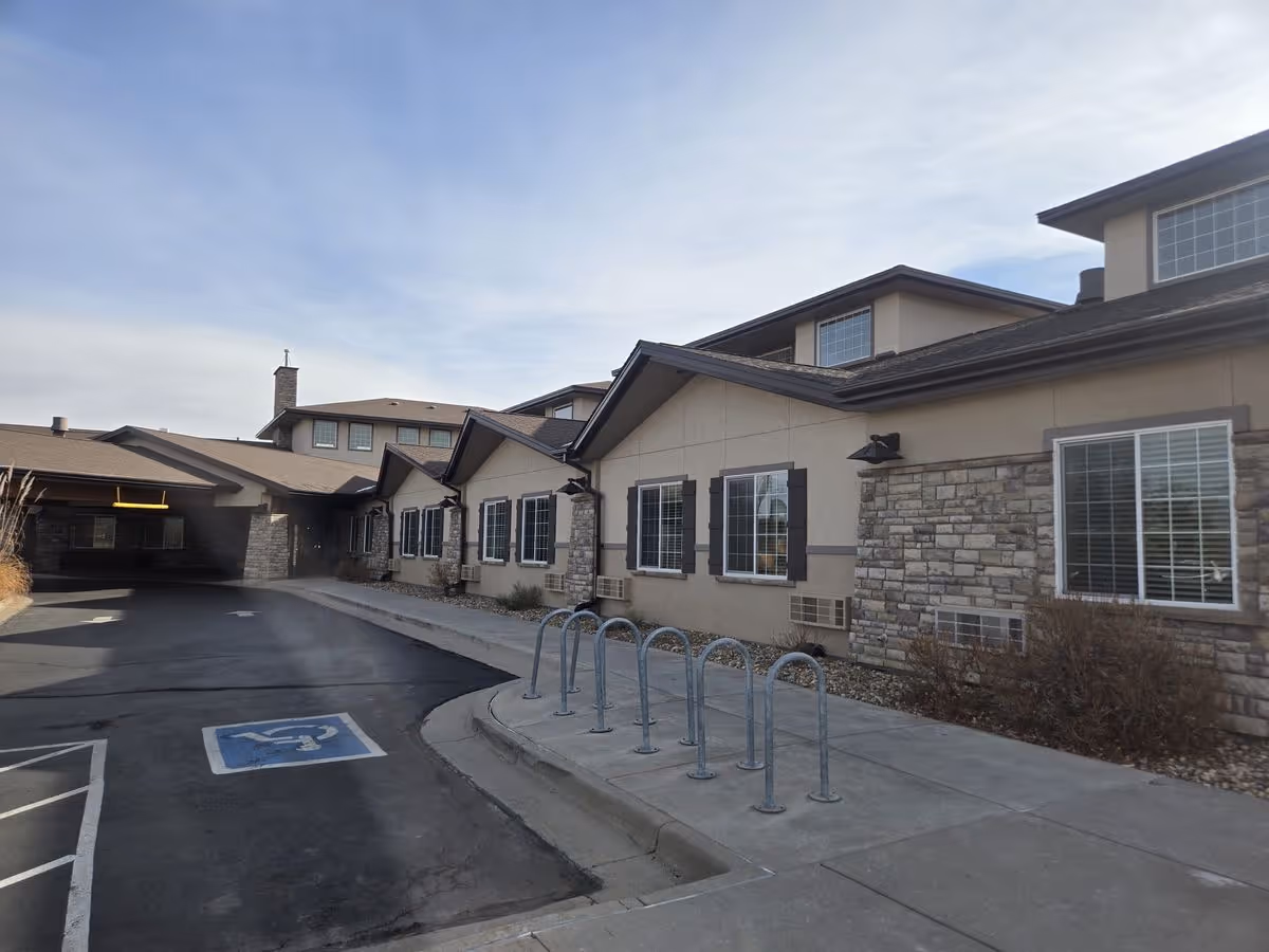 Front exterior of a single-story care facility with stone accents, multiple windows, a covered drop-off entrance, bike racks, and a marked handicap parking space.