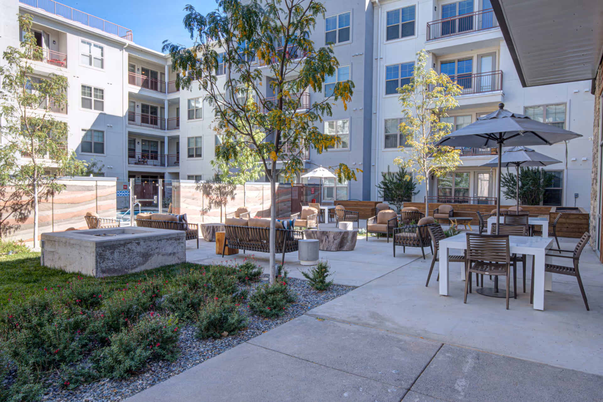 Outdoor courtyard area at Overture Albuquerque featuring seating arrangements with chairs, tables, umbrellas, and a fire pit surrounded by greenery and trees, with a multi-story residential building in the background.