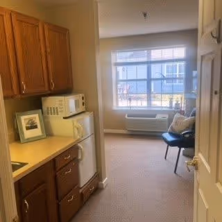 View of a small kitchen area with wooden cabinets, a microwave, and a mini refrigerator on the left side. Beyond the kitchen, there is a carpeted living space with a window, an air conditioning unit below the window, and a chair with cushions near the window.