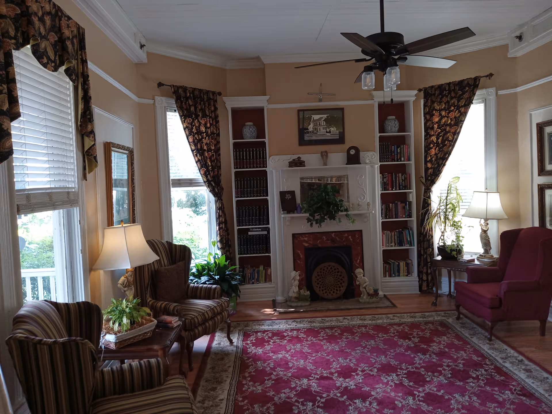 A cozy living room with a red patterned area rug, striped armchairs, a burgundy wingback chair, two table lamps, and a ceiling fan. The room features a white fireplace with decorative items and built-in bookshelves on either side filled with books and vases. Large windows with floral curtains allow natural light to enter.