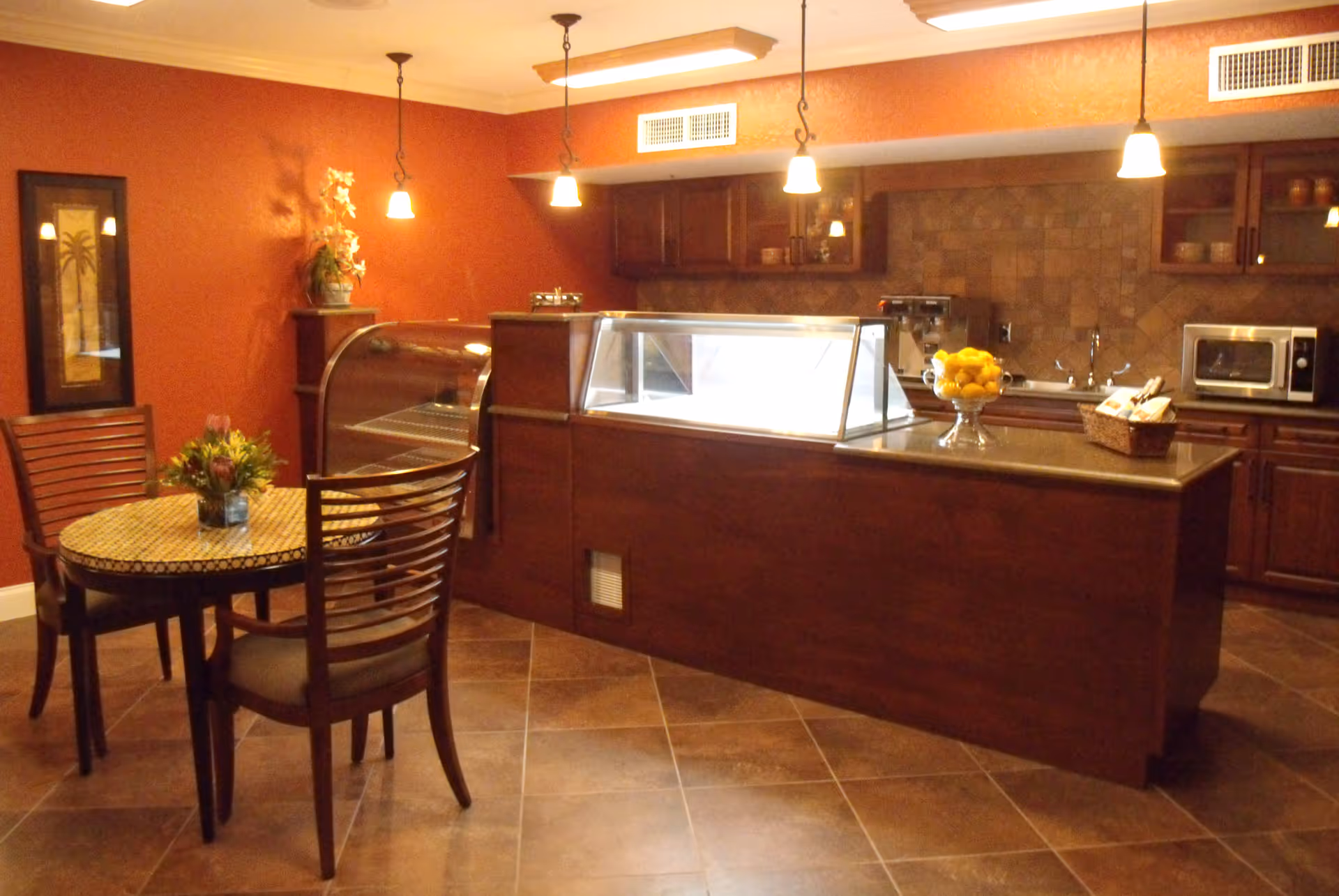 Interior view of a kitchen and dining area with warm lighting. The kitchen features dark wooden cabinets, a countertop with a glass display case, a microwave, and a bowl of lemons. There is a small round dining table with four chairs and a floral centerpiece. The walls are painted a warm reddish color, and the floor is tiled.