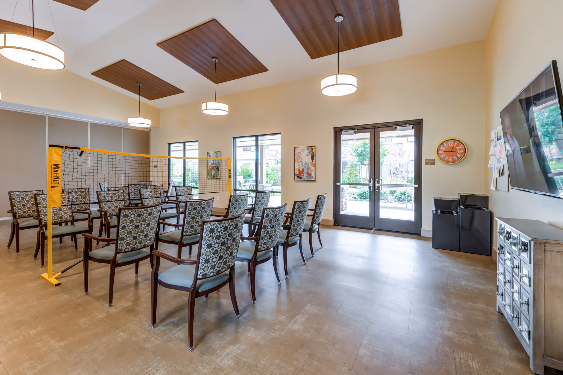A bright room with multiple chairs arranged in two rows facing each other with a mini volleyball net set up in the middle. The room has large windows and double glass doors leading outside. There are three ceiling lights, a wall clock, a mounted TV, and some artwork on the walls.