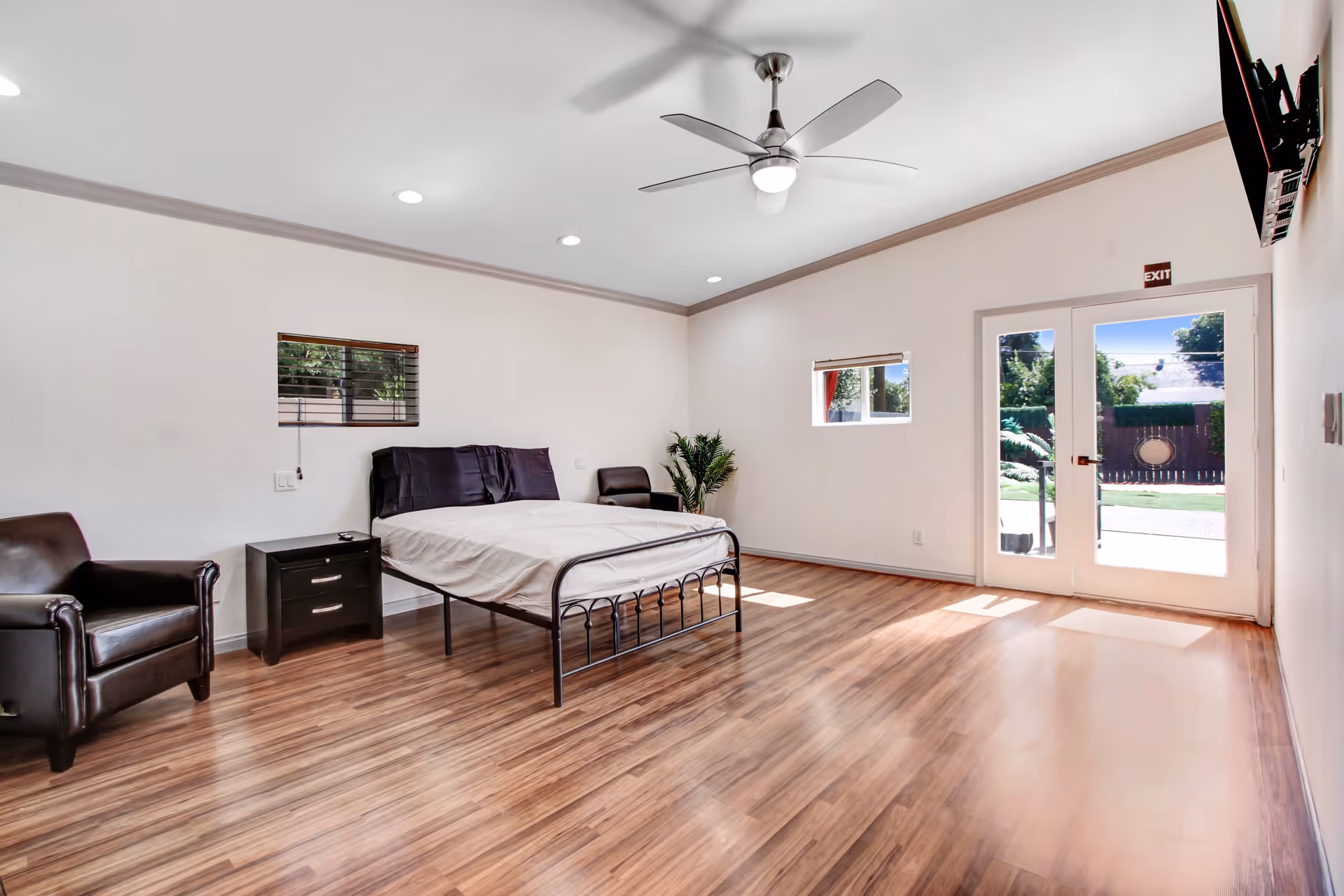 A spacious, bright bedroom with wooden flooring, a metal-framed bed with white bedding, two dark leather armchairs, a small black nightstand, a ceiling fan with light, two small windows, and glass double doors leading outside to a garden area.