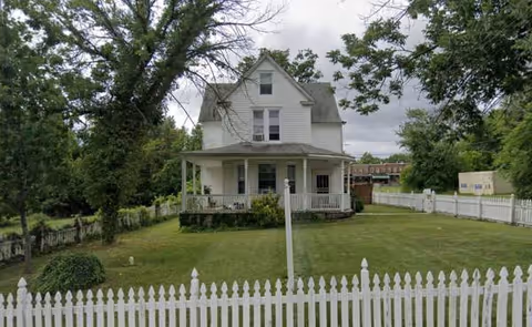 A white two-story house with a porch, fenced yard, and tall trees.