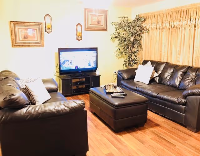 A cozy living room with two black leather sofas facing each other, a black ottoman in the center, a flat-screen TV on a black TV stand, framed artwork and wall sconces on the wall, a large potted plant in the corner, and beige curtains covering the window.