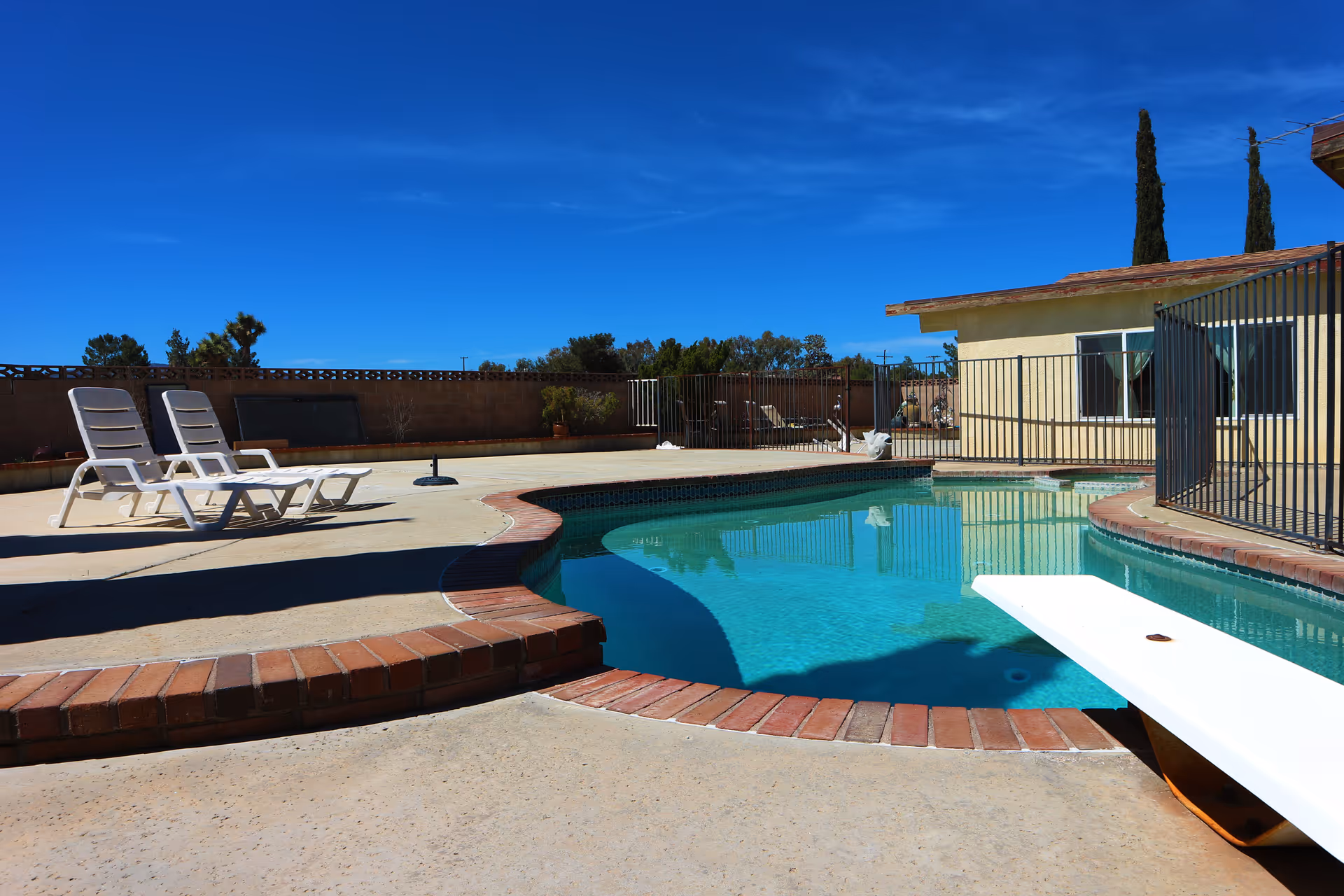 Outdoor swimming pool with a diving board and two white lounge chairs on the pool deck under a clear blue sky. The pool area is enclosed by a fence and a low brick wall, with a single-story building visible in the background.