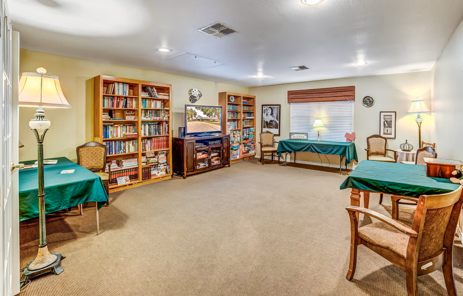 A spacious communal living/activity room with bookshelves, a TV, and multiple tables and chairs covered with green tablecloths.