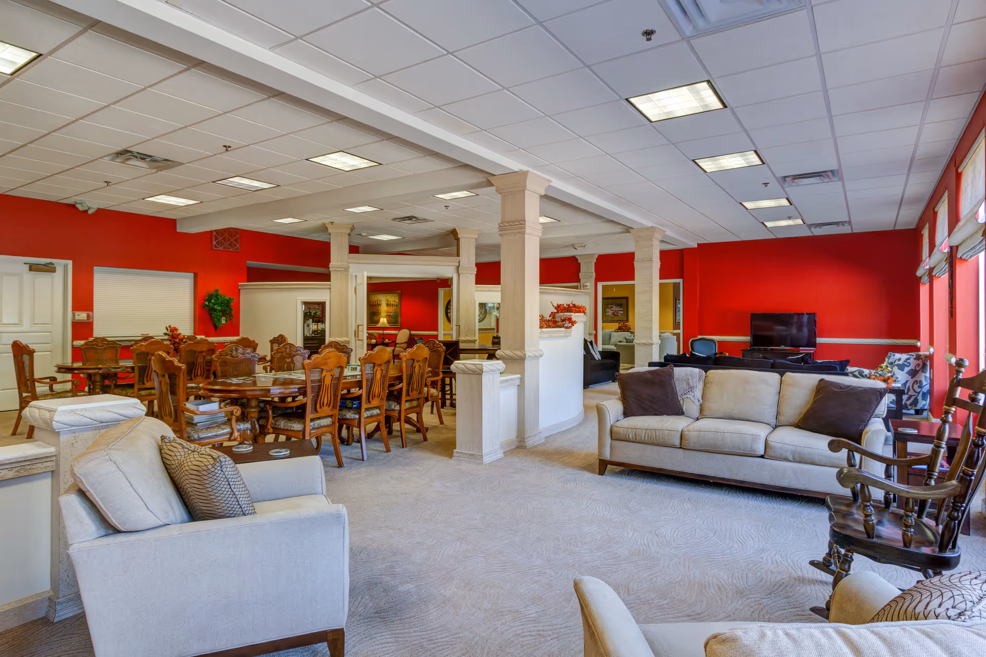 Spacious common area with beige sofas, armchairs, and wooden rocking chairs arranged around a television. The room features red walls, large windows with blinds, and a dining area with multiple wooden tables and chairs. Decorative columns and a carpeted floor complete the cozy, inviting atmosphere.