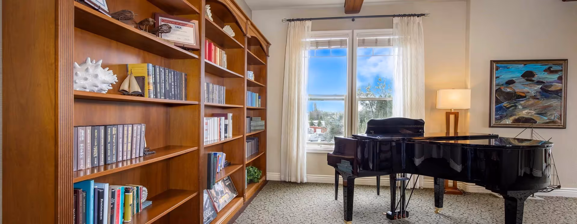 A bright room with large wooden bookshelves filled with books and decorative items on the left, a black grand piano in the center near a window with white curtains, a floor lamp with a rectangular shade, and a framed painting of rocks in water on the wall.
