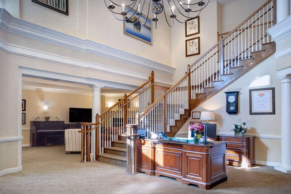 Interior view of a senior living facility lobby with a wooden reception desk adorned with a vase of flowers, a staircase with wooden handrails and white balusters leading to an upper floor, a wall clock, framed certificates, and artwork on the walls. In the background, there is a sitting area with a piano and a television.