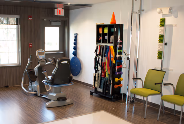 Bright exercise room featuring a recumbent exercise bike, a rack of colorful dumbbells and resistance bands, and green chairs.