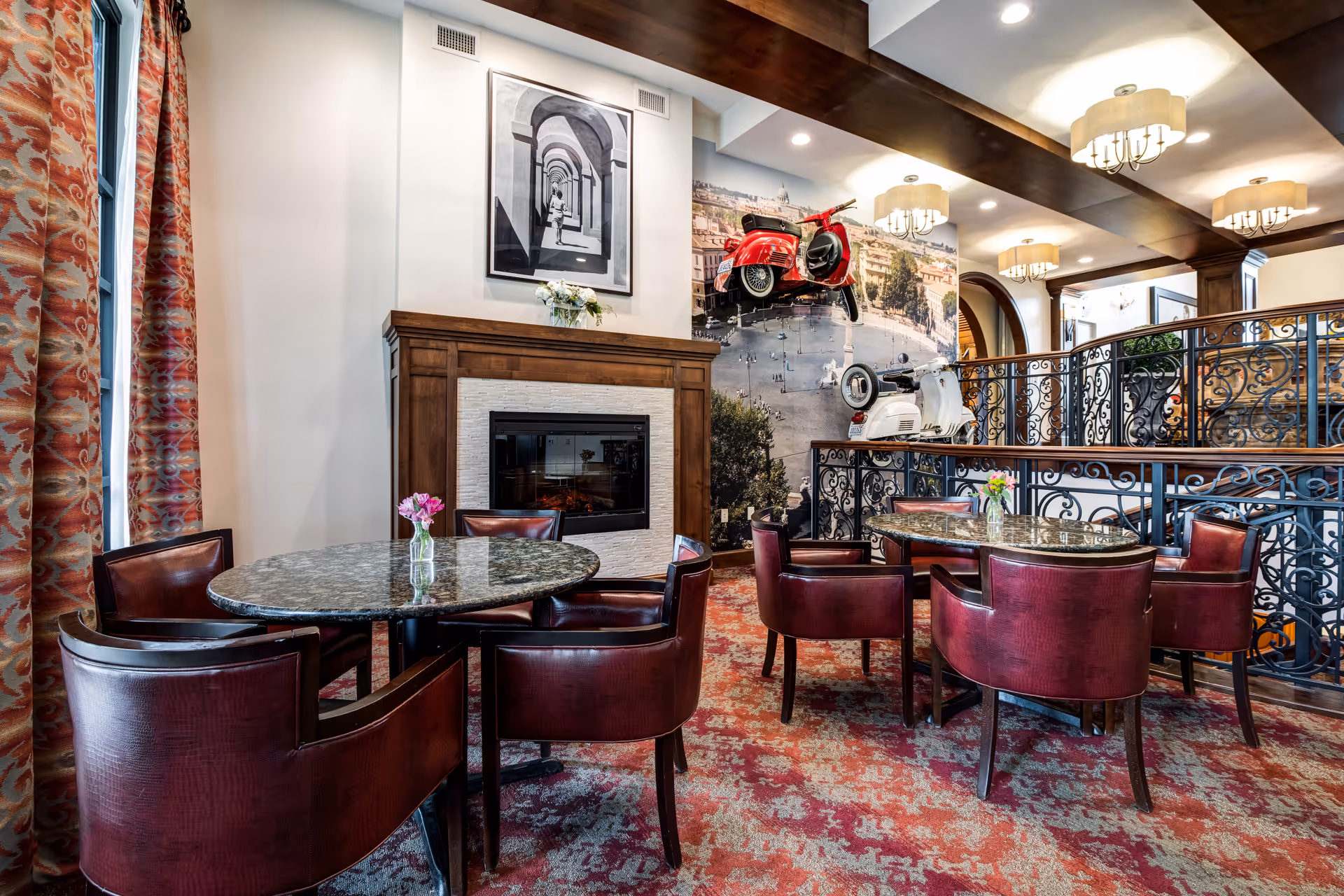 A cozy interior seating area with round marble tables and red leather chairs on a patterned red carpet. There is a wooden fireplace with a black and white framed photo above it. The wall behind features a large mural of a city square with two vintage scooters mounted on it. The ceiling has wooden beams and modern light fixtures, and decorative wrought iron railings separate the seating area from another level.