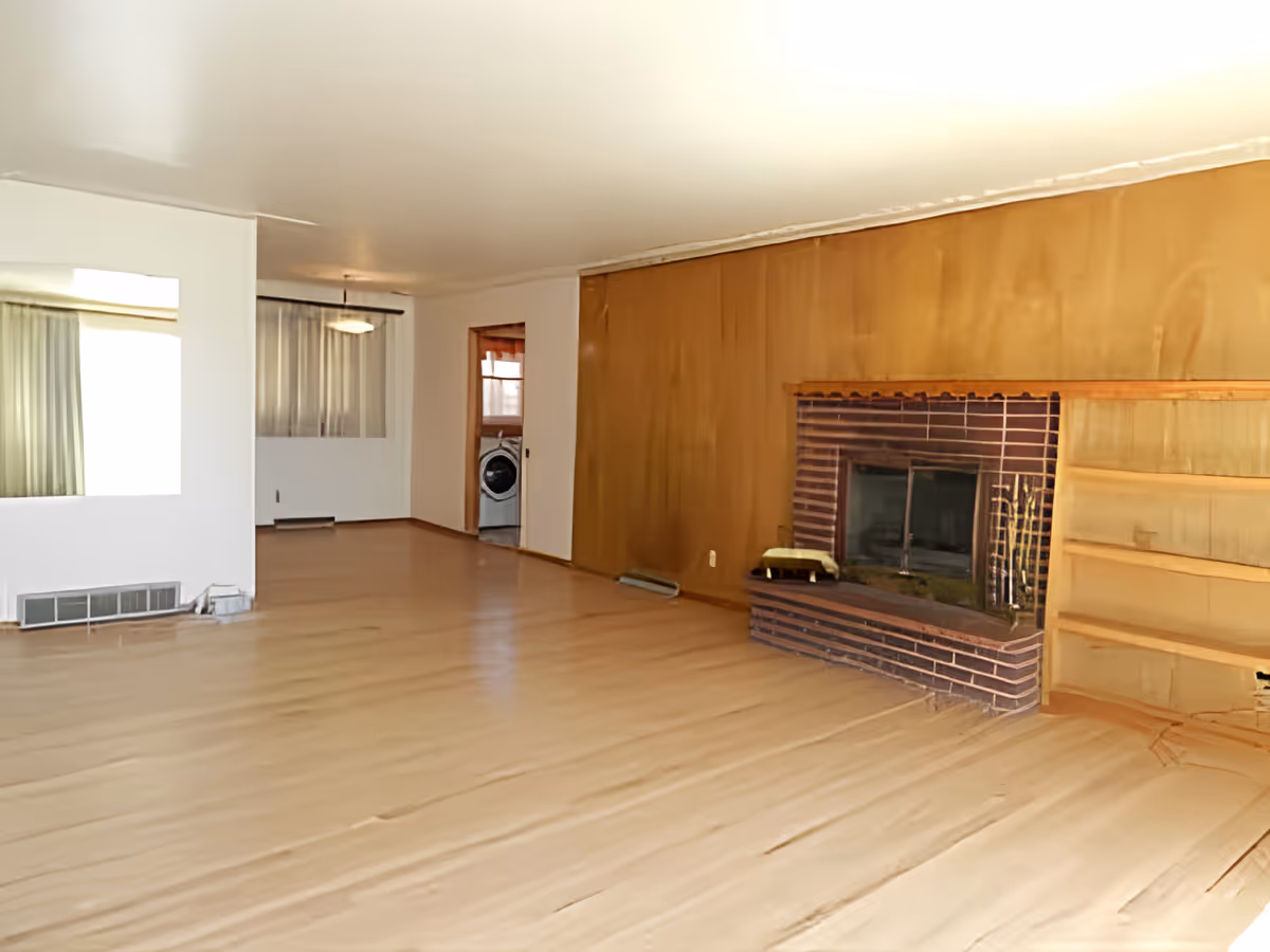 Empty living room with light wood flooring, a brick fireplace with a wooden mantel on the right wall, built-in wooden shelves next to the fireplace, and a large window with curtains in the background. There is an open doorway leading to another room with a visible washing machine.