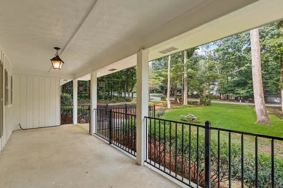 Covered outdoor porch area with white ceiling and columns, black metal railing, and a hanging lantern light fixture. The porch overlooks a green lawn with trees and shrubs in the background.