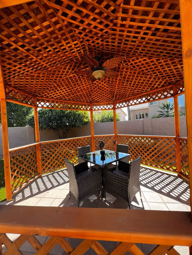 A wooden gazebo with lattice walls and a ceiling fan, containing a glass-top table surrounded by four wicker chairs, situated outdoors with a view of a garden and neighboring houses.
