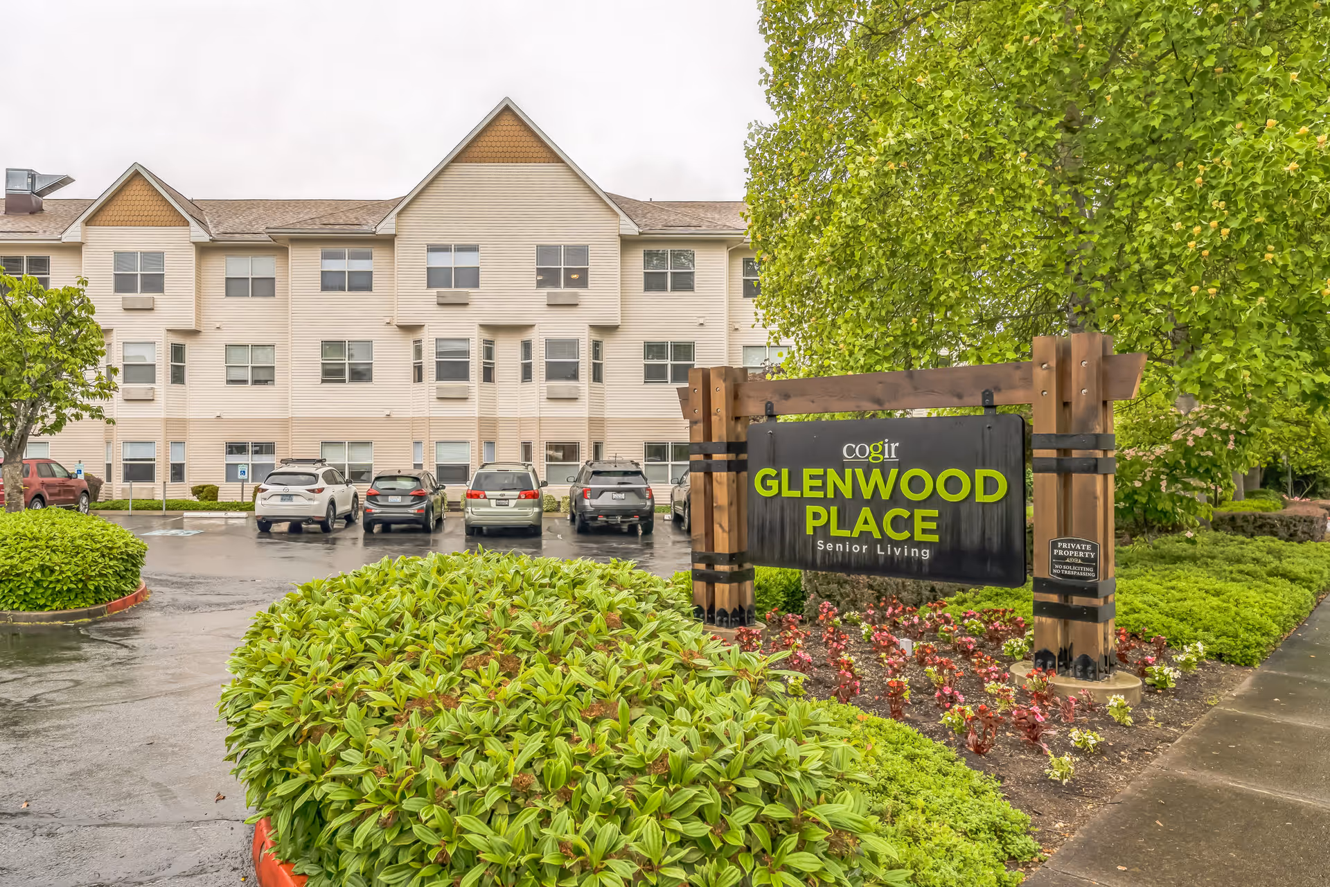 Exterior view of Cogir Glenwood Place senior living facility showing a three-story beige building with multiple windows, a parking lot with several cars, and a large wooden sign with green and white text reading 'Cogir Glenwood Place Senior Living' surrounded by green bushes and trees on a cloudy day.