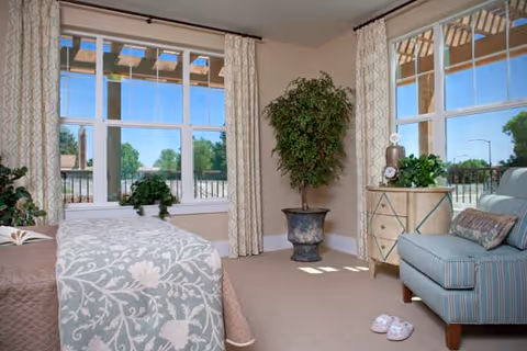 A bright and cozy bedroom with large windows letting in natural light. The room features a bed with a floral patterned bedspread, a striped armchair with a decorative pillow, a small chest of drawers with plants and decor on top, and a large potted plant in the corner. Light-colored curtains frame the windows, and a pair of slippers is placed on the carpeted floor.
