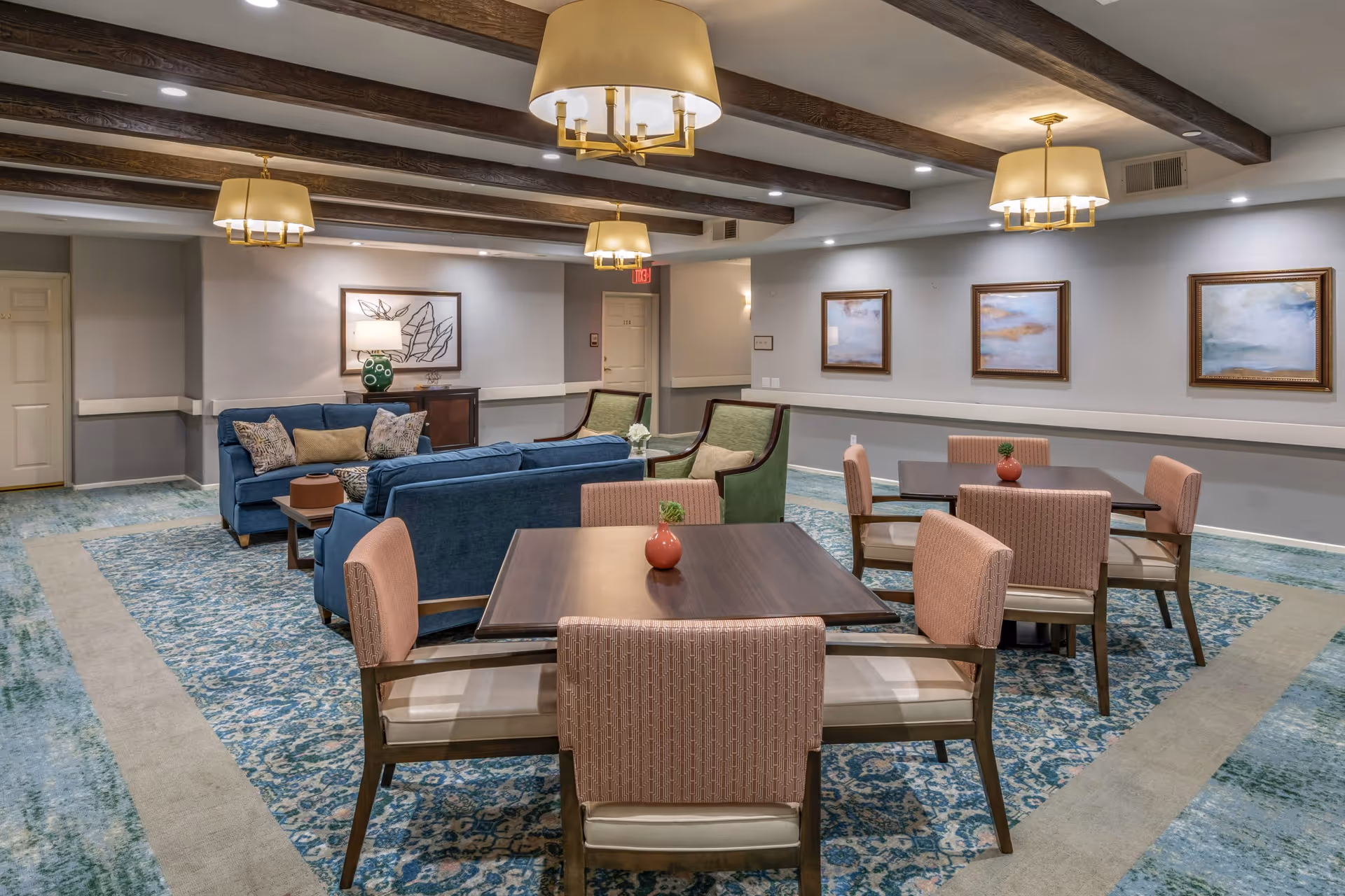 A spacious and well-lit common area featuring two square wooden tables each surrounded by four upholstered chairs with pinkish fabric. Behind the tables, there are two blue sofas and two green armchairs arranged around a small coffee table. The room has a patterned blue and beige carpet, wooden ceiling beams, and three large pendant lights with beige lampshades. The walls are decorated with framed artwork and a table with a lamp and decorative items is visible in the background.