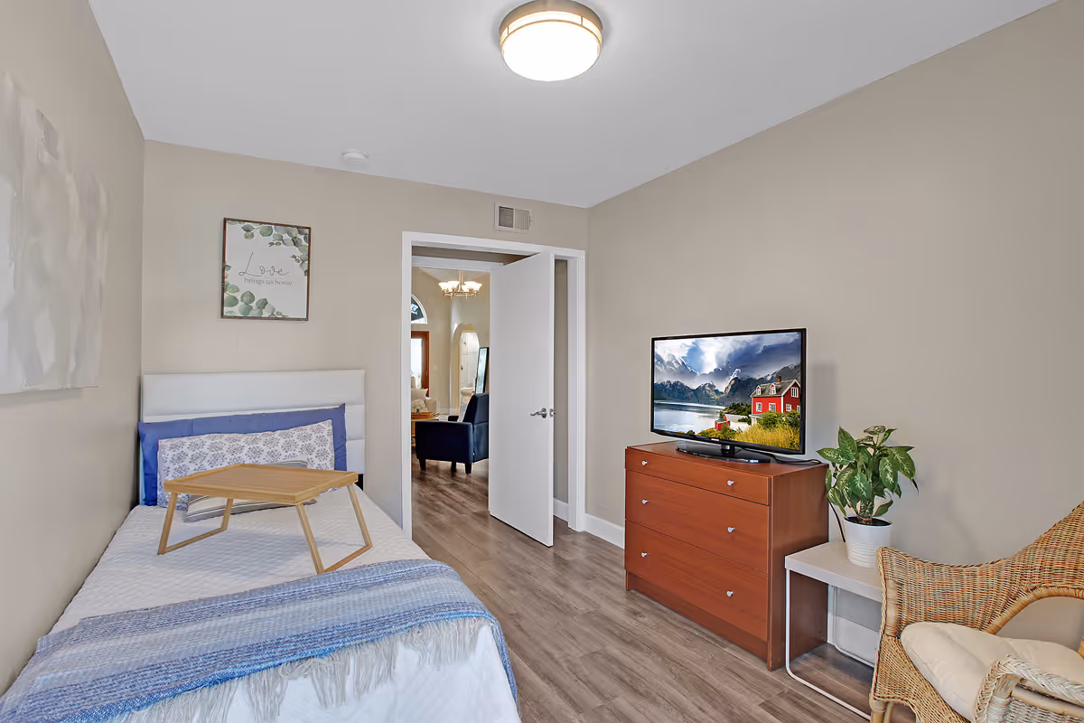 A small bedroom in a senior living facility with a single bed covered with white and blue bedding, a wooden tray on the bed, a wooden dresser with a flat-screen TV displaying a scenic landscape, a wicker chair with a cushion, a potted plant on a small white table, and an open door leading to another room with a chandelier and blue armchairs.