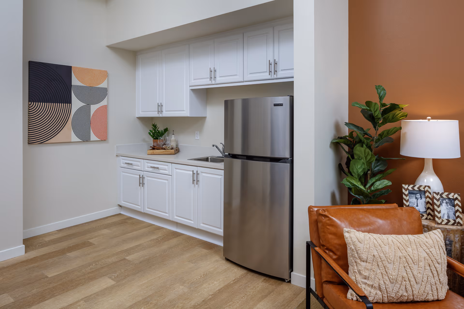 A small kitchen area with white cabinets, a stainless steel refrigerator, and a sink. To the right, there is a brown leather chair with a textured beige pillow, a green plant, a white table lamp, and two framed black and white photos on a wooden side table. On the left wall, there is a colorful abstract artwork with circular patterns.