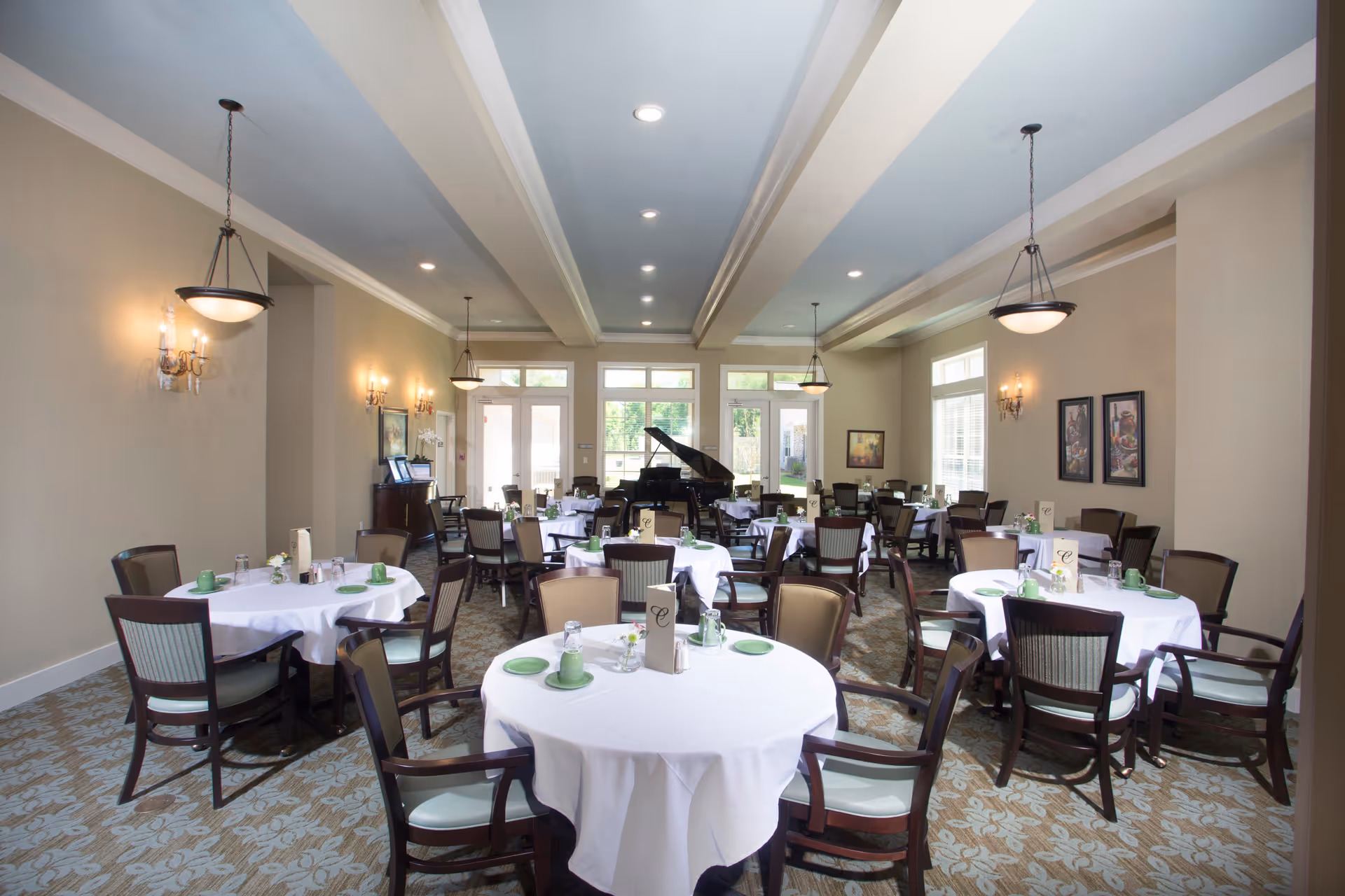 A spacious dining room with multiple round tables covered with white tablecloths, each set with green plates, cups, and glassware. The room features beige walls, patterned carpet, hanging pendant lights, wall sconces, and large windows letting in natural light. A grand piano is visible at the far end of the room.
