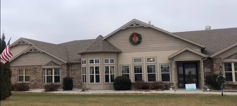 Front exterior of a single-story brick and siding senior care cottage with multiple windows, an American flag, and a wreath above the entrance.