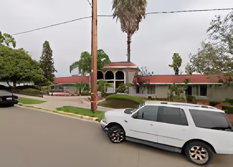 Exterior view of a single-story building with a red roof and arched windows, surrounded by trees and greenery. A white station wagon is parked on the street in front of the building under an overcast sky.