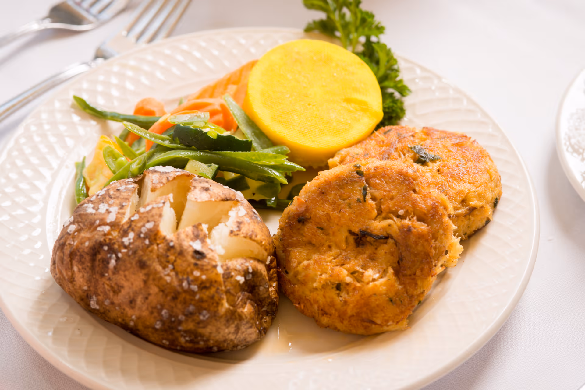 A plated meal featuring a baked potato, two crab cakes, mixed vegetables and a yellow side on a white plate.