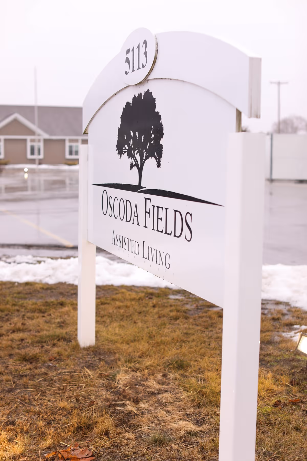 A white outdoor sign for Oscoda Fields Assisted Living with the number 5113 displayed at the top. The sign features a black silhouette of a tree and is set on a patch of grass with some snow and a parking lot in the background.